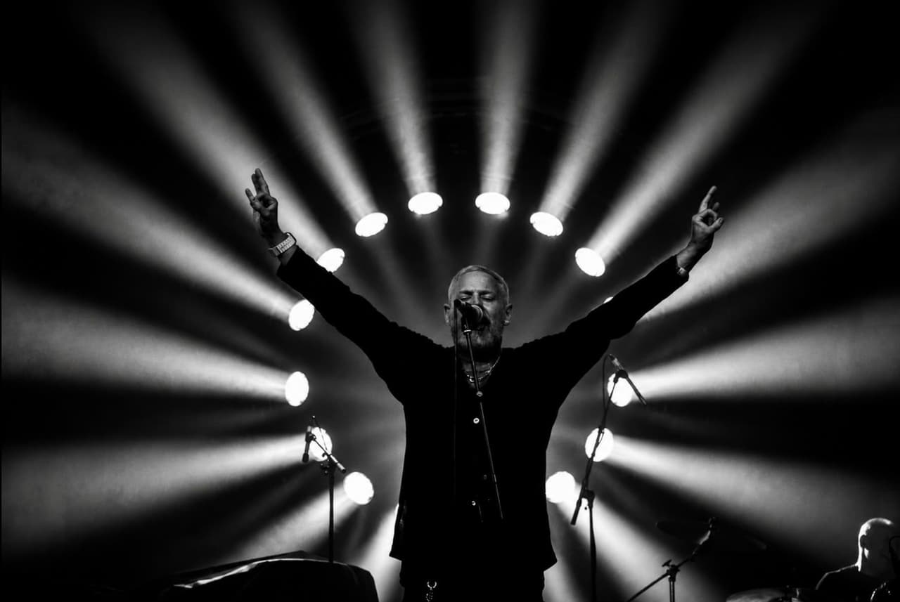 Black and white: a frontman with both arms raised inside a ring of stage lights.