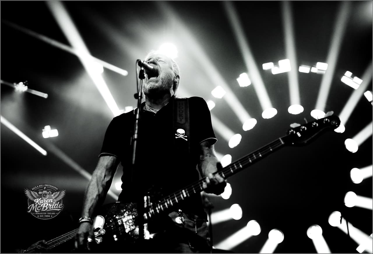 Black and white: Peter Hook sings with his bass slung low, stage lights fanned out behind him.