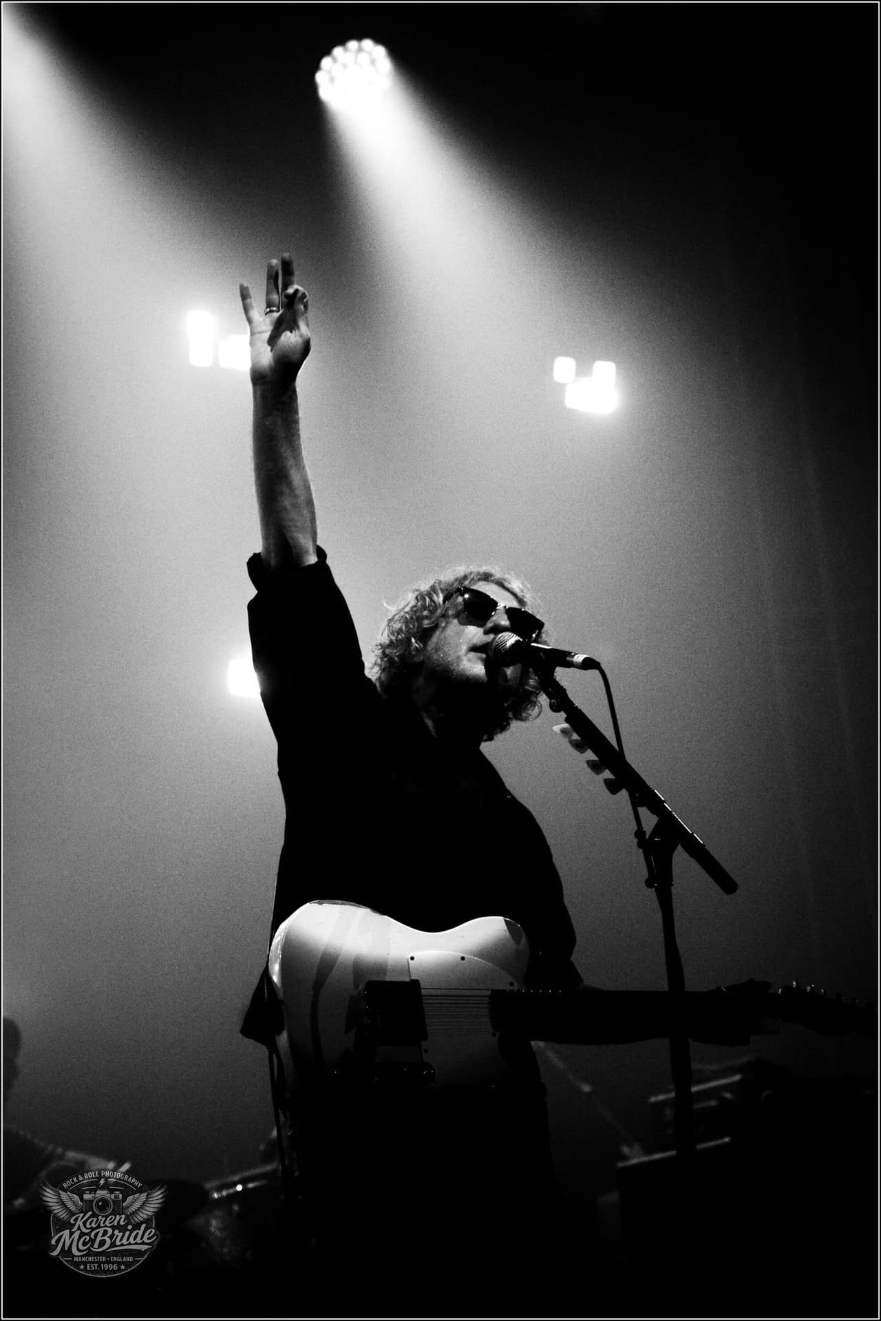 Black and white: a singer in sunglasses raises two fingers to the lights, white Telecaster at his waist.