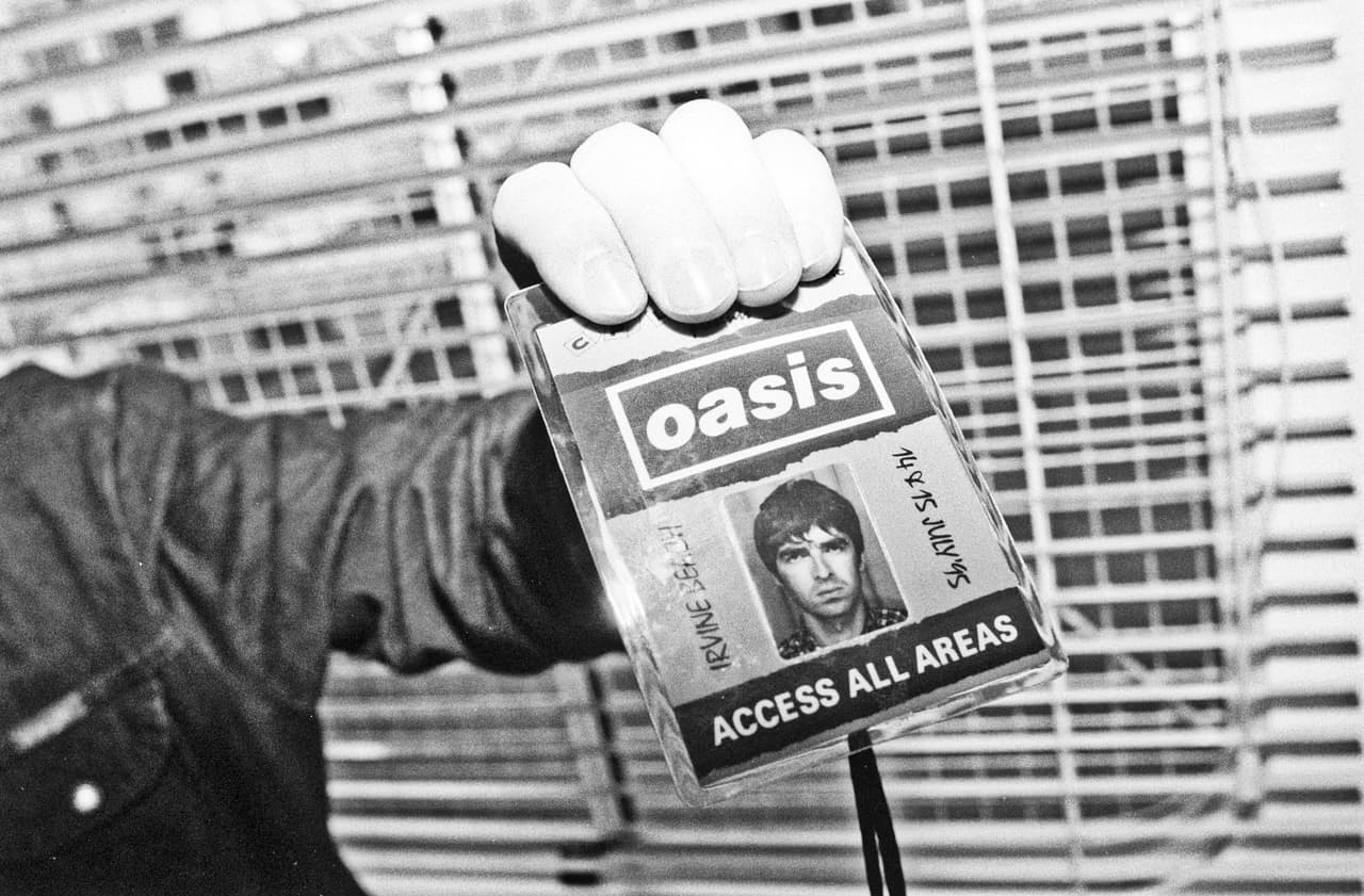 Black and white: a hand holds up an Oasis Access All Areas backstage pass carrying Noel Gallagher's photo.