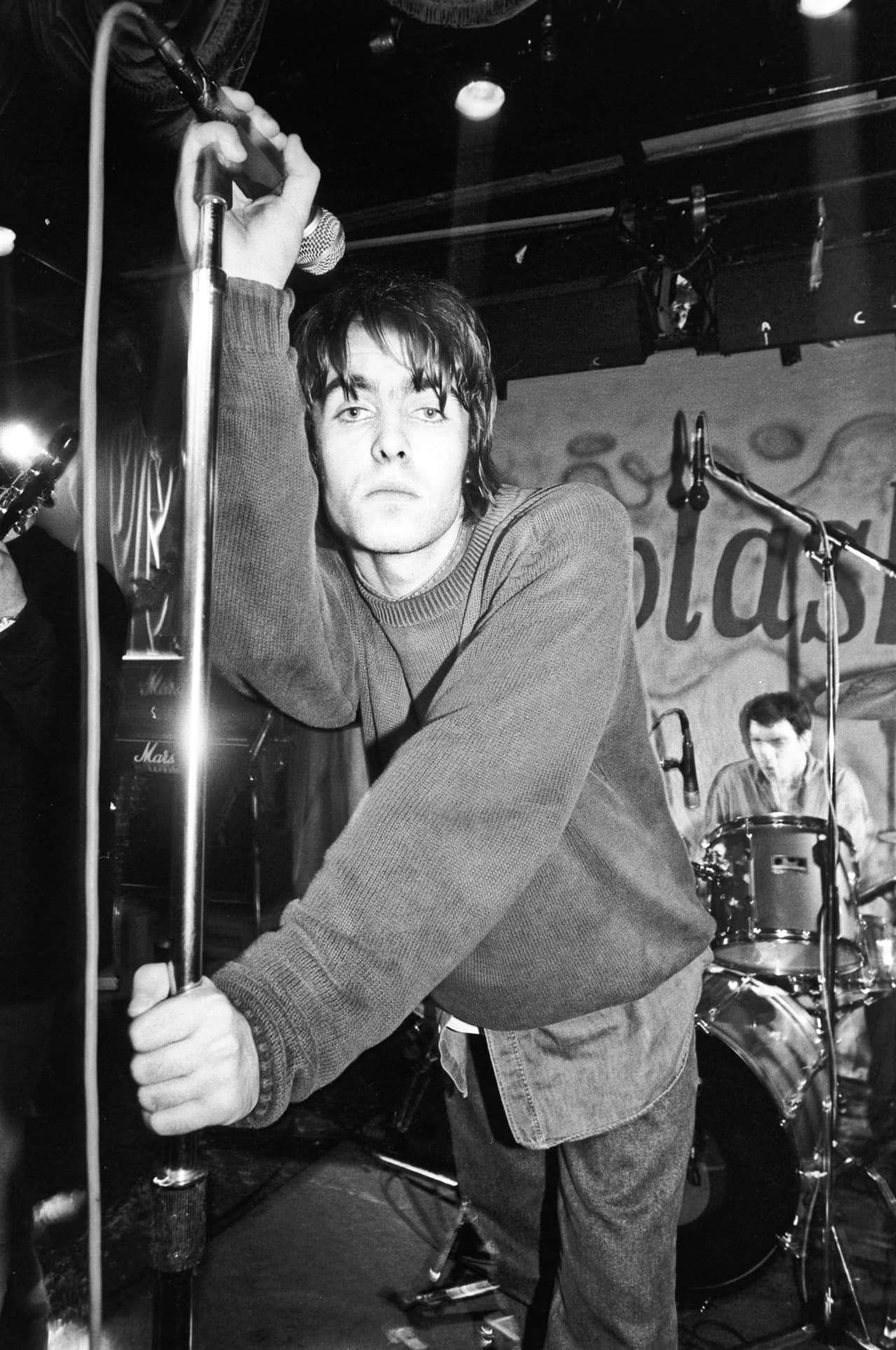 Liam Gallagher leans over the mic stand and eyes the camera, drummer behind, black and white.