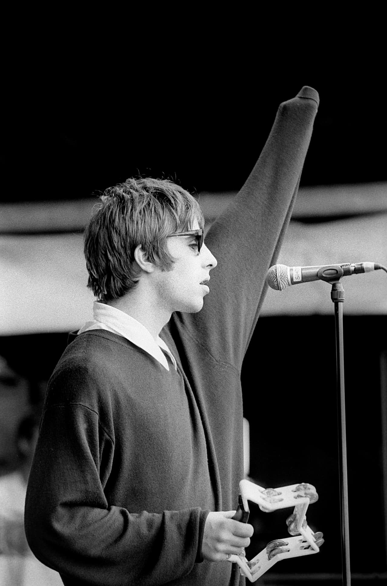 Liam Gallagher in shades at an outdoor mic, arm aloft, star tambourine in hand, black and white.