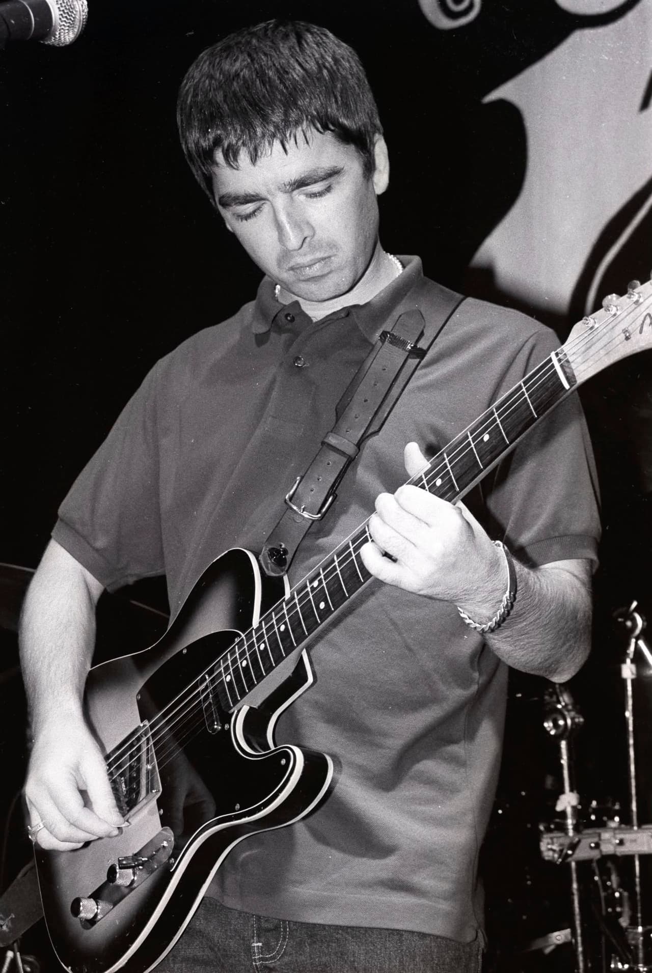 Noel Gallagher head down over his guitar in a polo shirt, black and white.