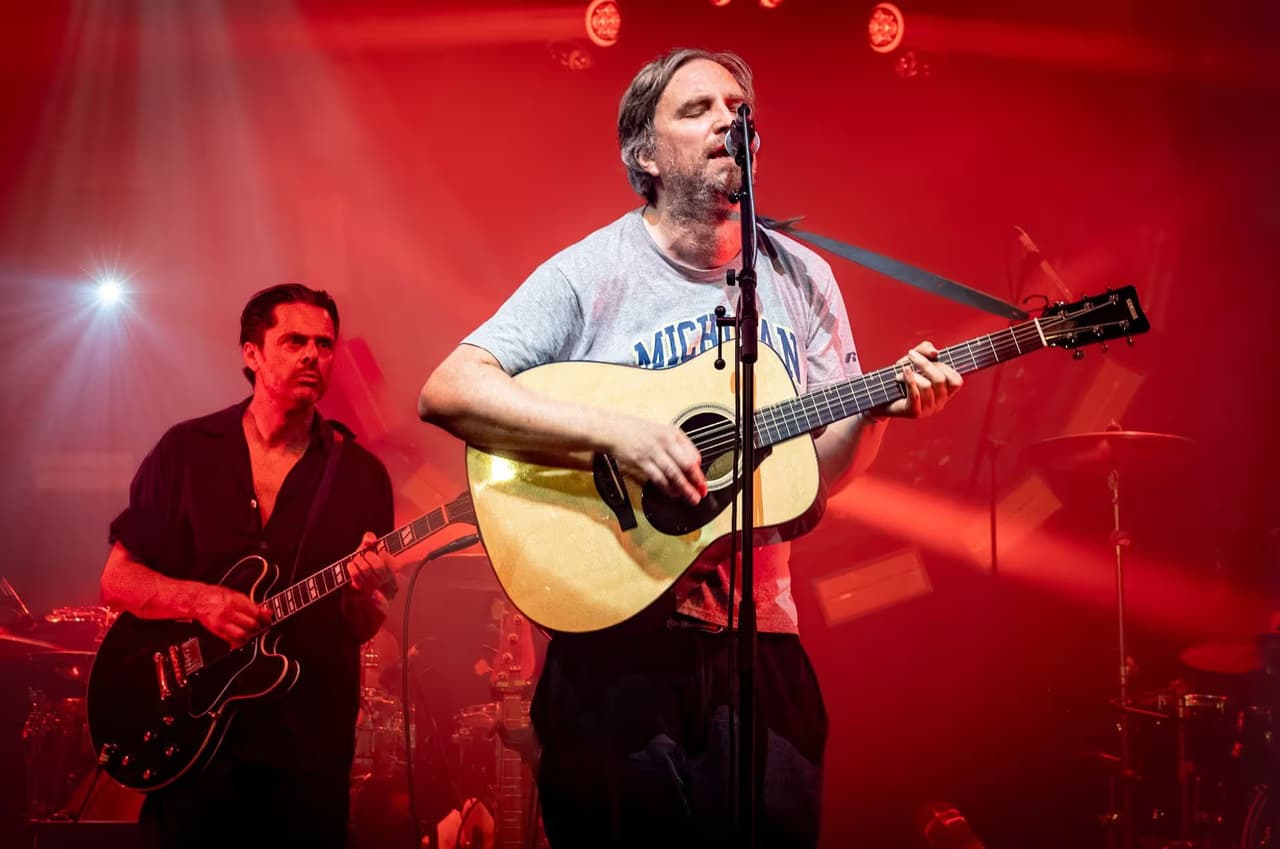An acoustic guitarist sings beside an electric guitarist, the stage bathed in red.