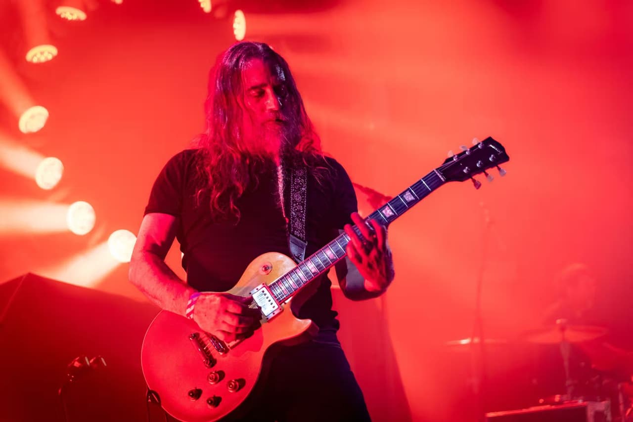 A long-haired guitarist plays a gold-top Les Paul in deep red light.