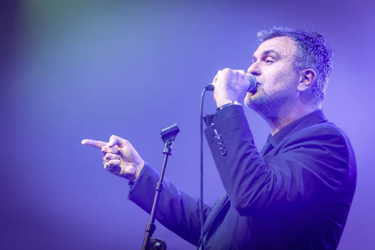 A singer in a dark suit lit blue and purple, pointing across the stage.