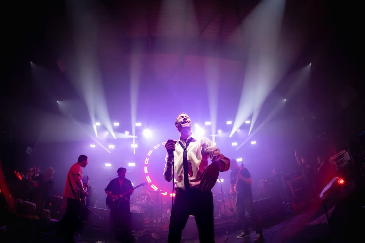 A wide stage shot in purple haze, the frontman lit at the centre of the beams.