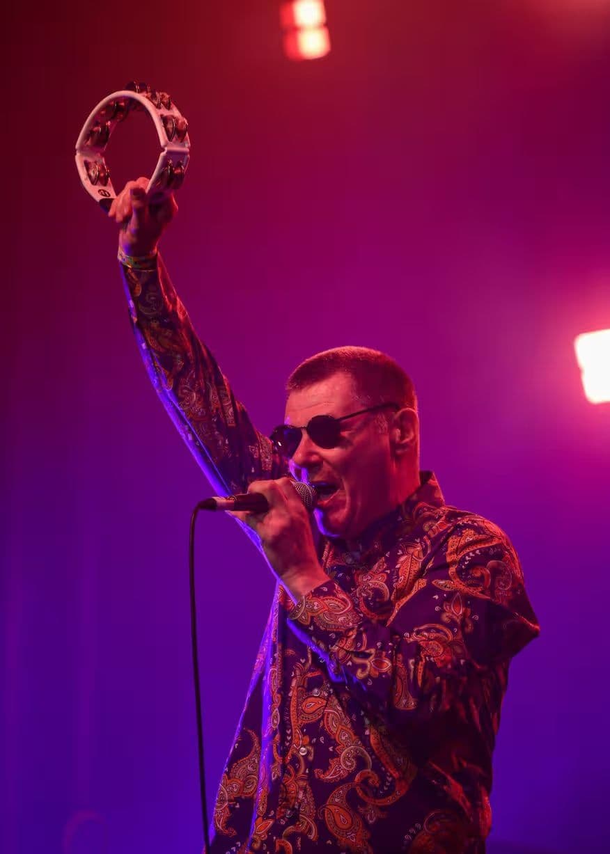 A singer in a paisley shirt raises a tambourine under purple and red light.