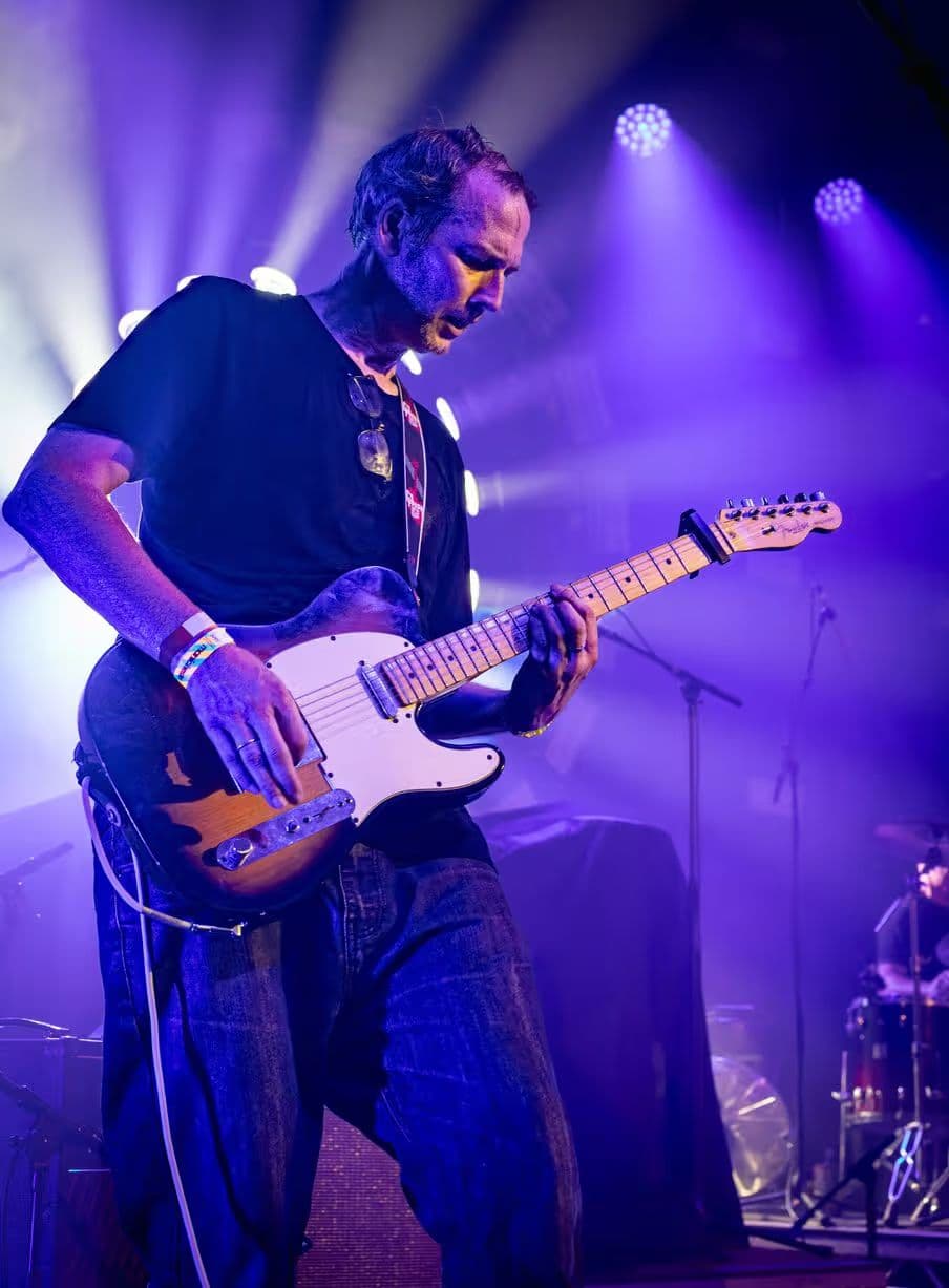 A guitarist plays a Telecaster under purple light, sunglasses hanging from his shirt.