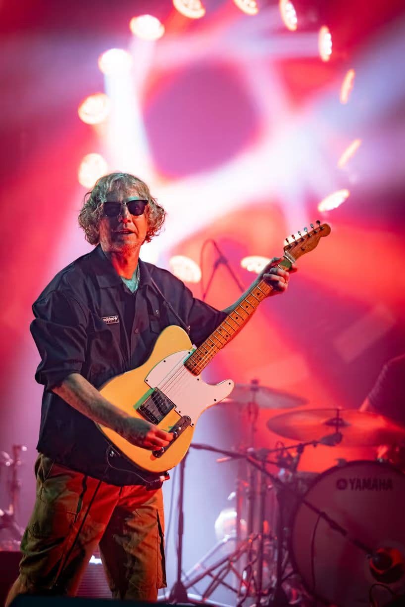 A guitarist in sunglasses with a blonde Telecaster, drum kit behind him in pink light.