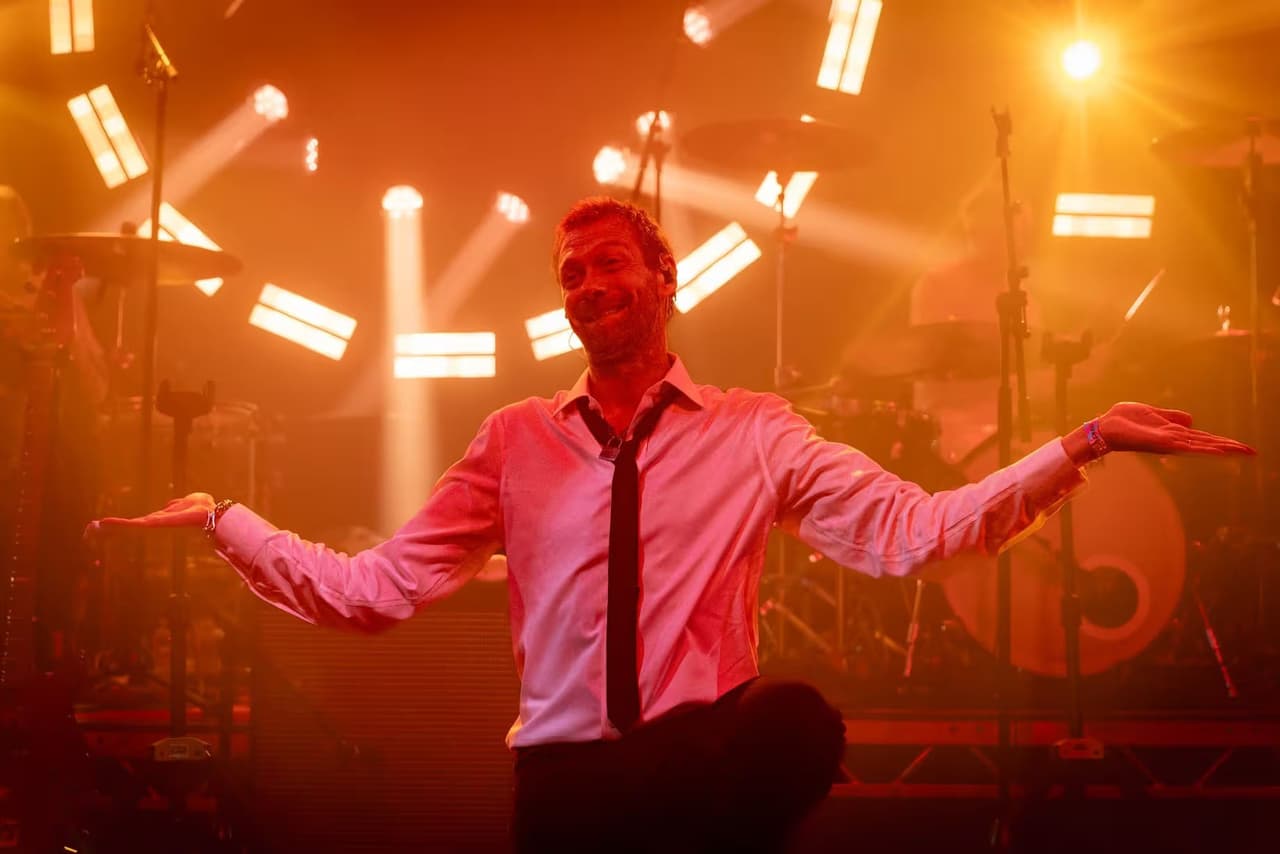 A frontman in a white shirt and black tie, arms out under orange stage light.