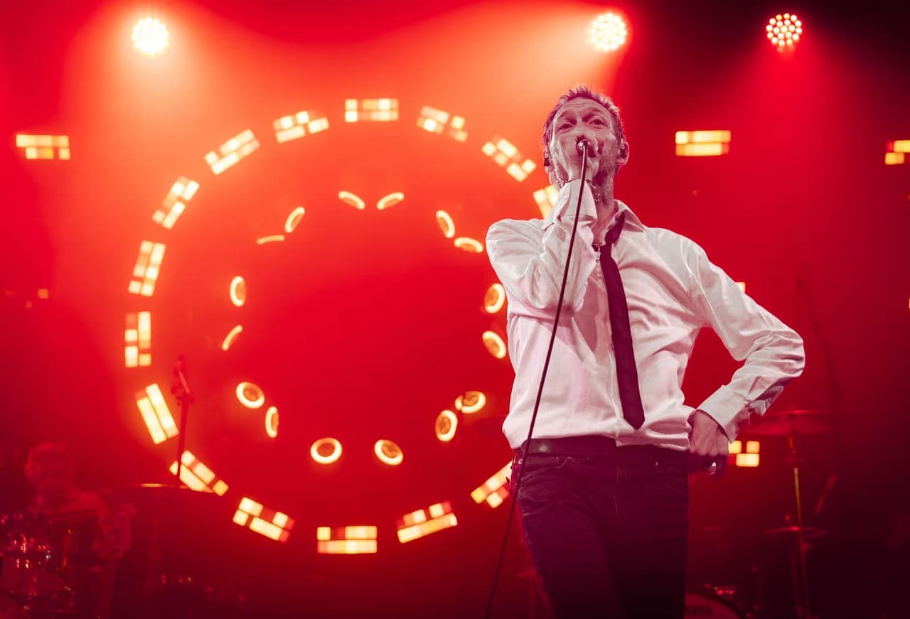 A frontman in a white shirt and black tie sings in front of a ring of red lights.